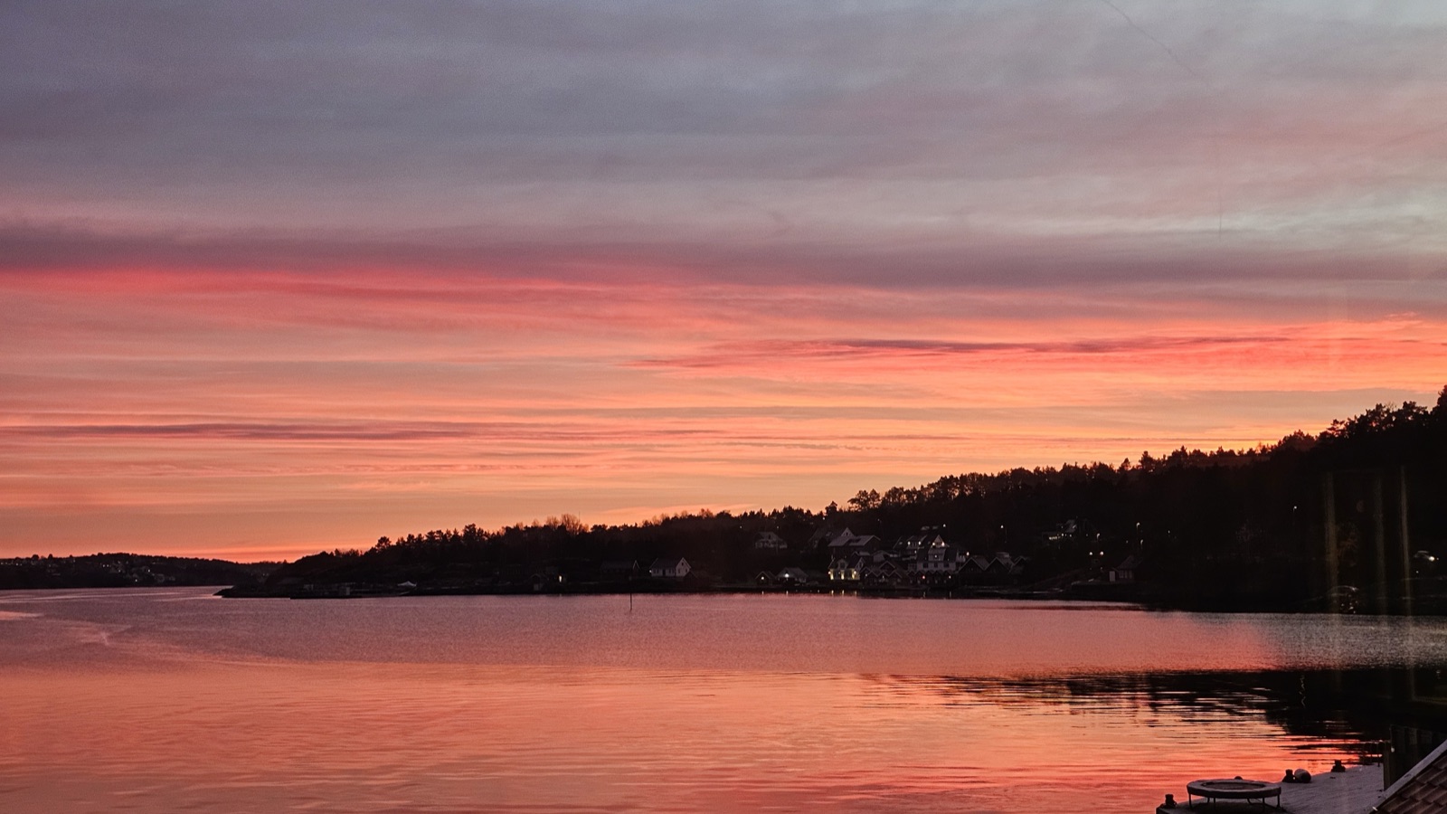 A pink and orange sunset over a calm bay, with lit windows along the darkening shoreline